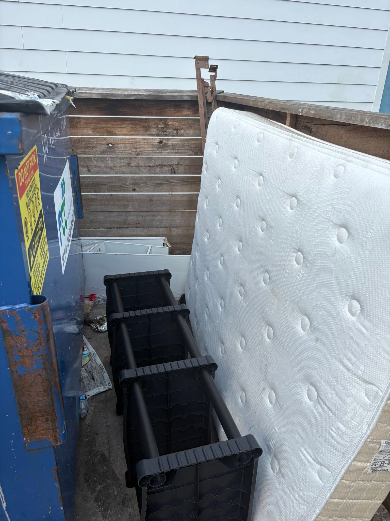 Outdoor yard with a black metal frame beside a large white mattress leaning against a wall.