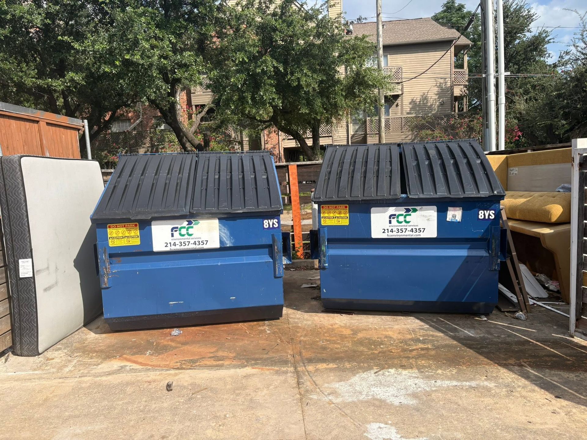 Two large blue dumpsters with black lids in a parking lot beside a wall and trees.
