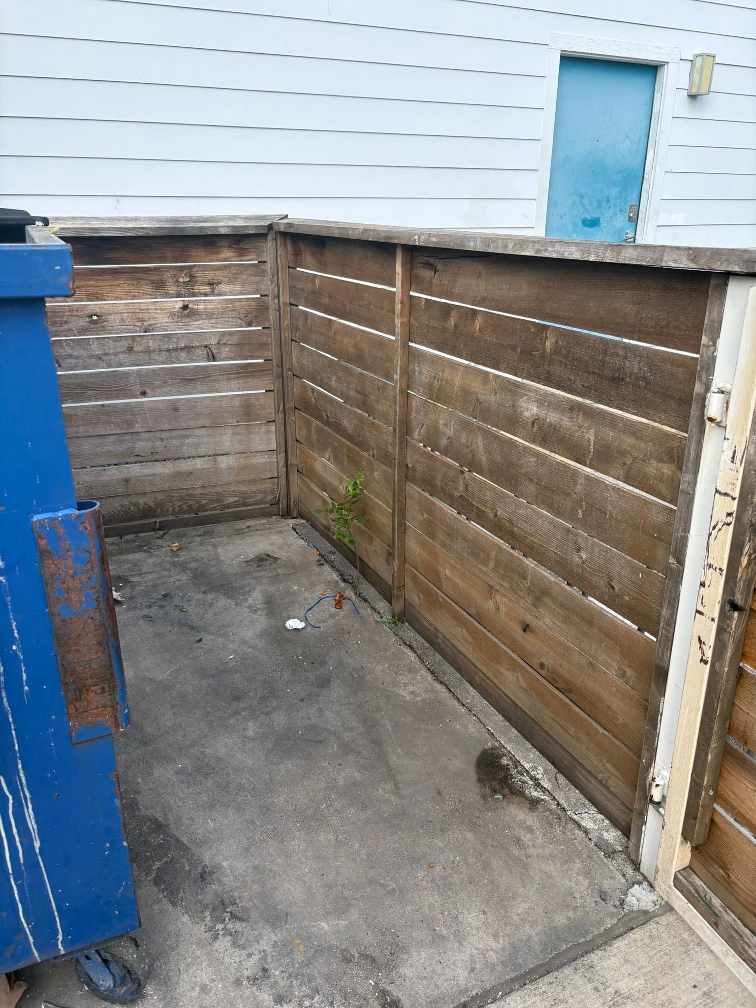 Small fenced concrete corner beside a white building, with a blue door and blue dumpster.