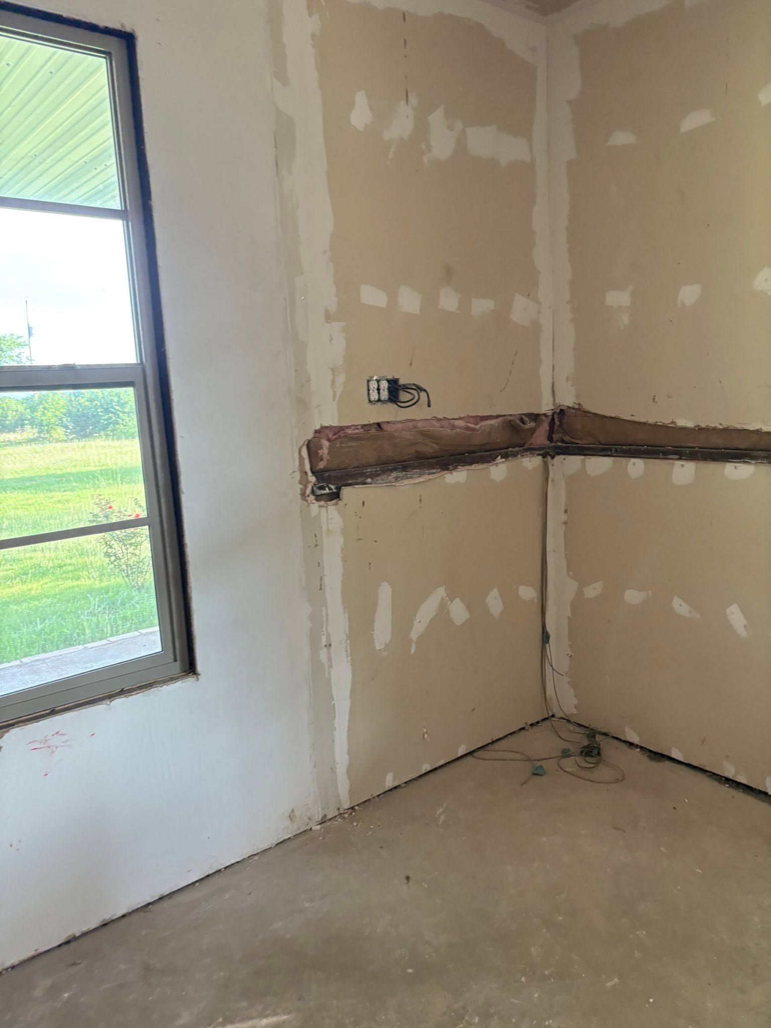 Empty unfinished room with drywall patches, a window on the left, and a wall-mounted work ledge or bracket.