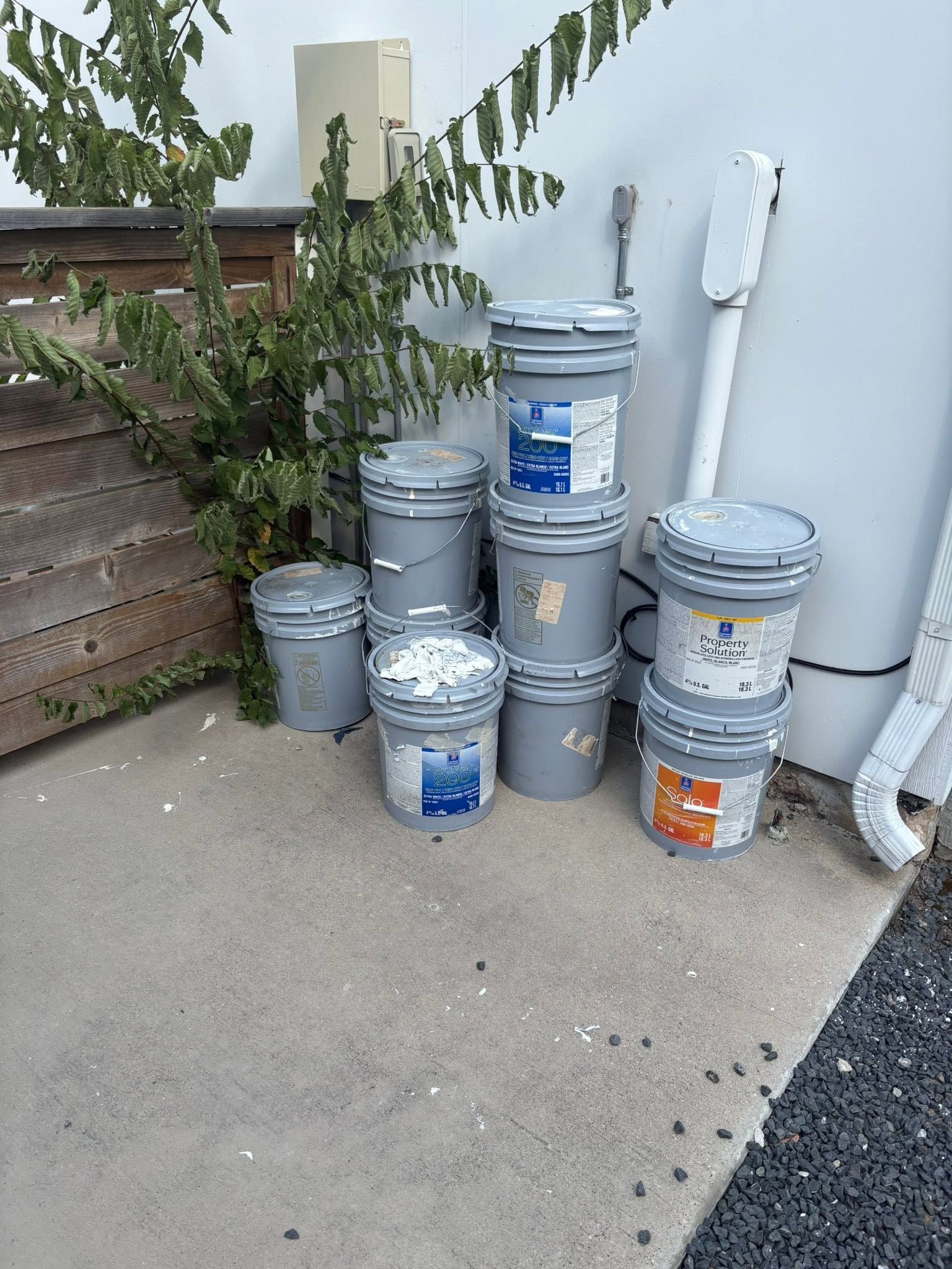 Stacked gray and blue paint buckets beside a house wall on a concrete patio, near a fence and greenery