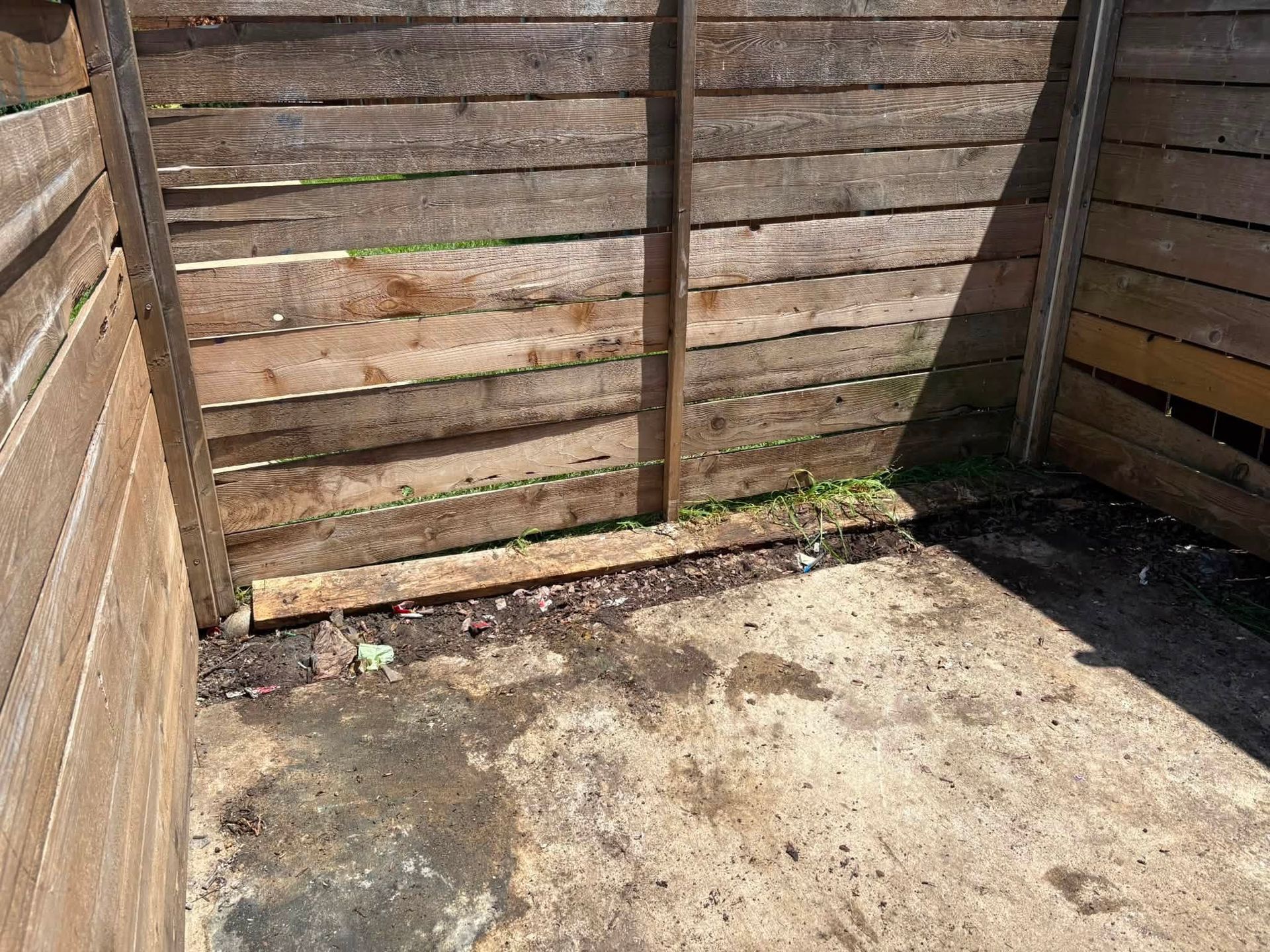 Empty weathered wooden corner with a dirty concrete floor and a small patch of weeds.