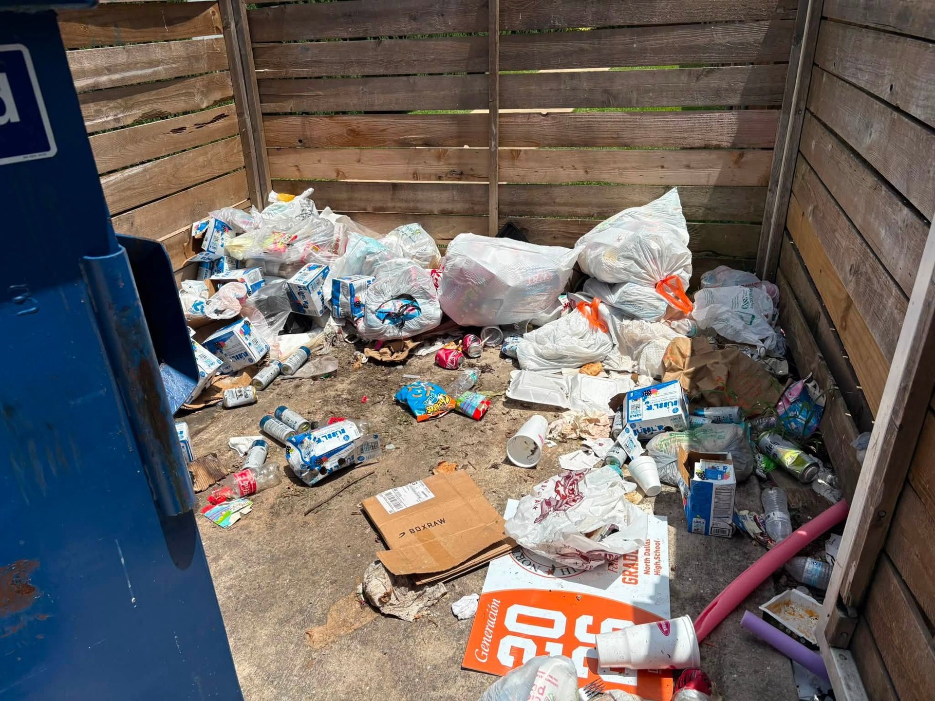 Overflowing trash and scattered cardboard fill a wooden dumpster enclosure.
