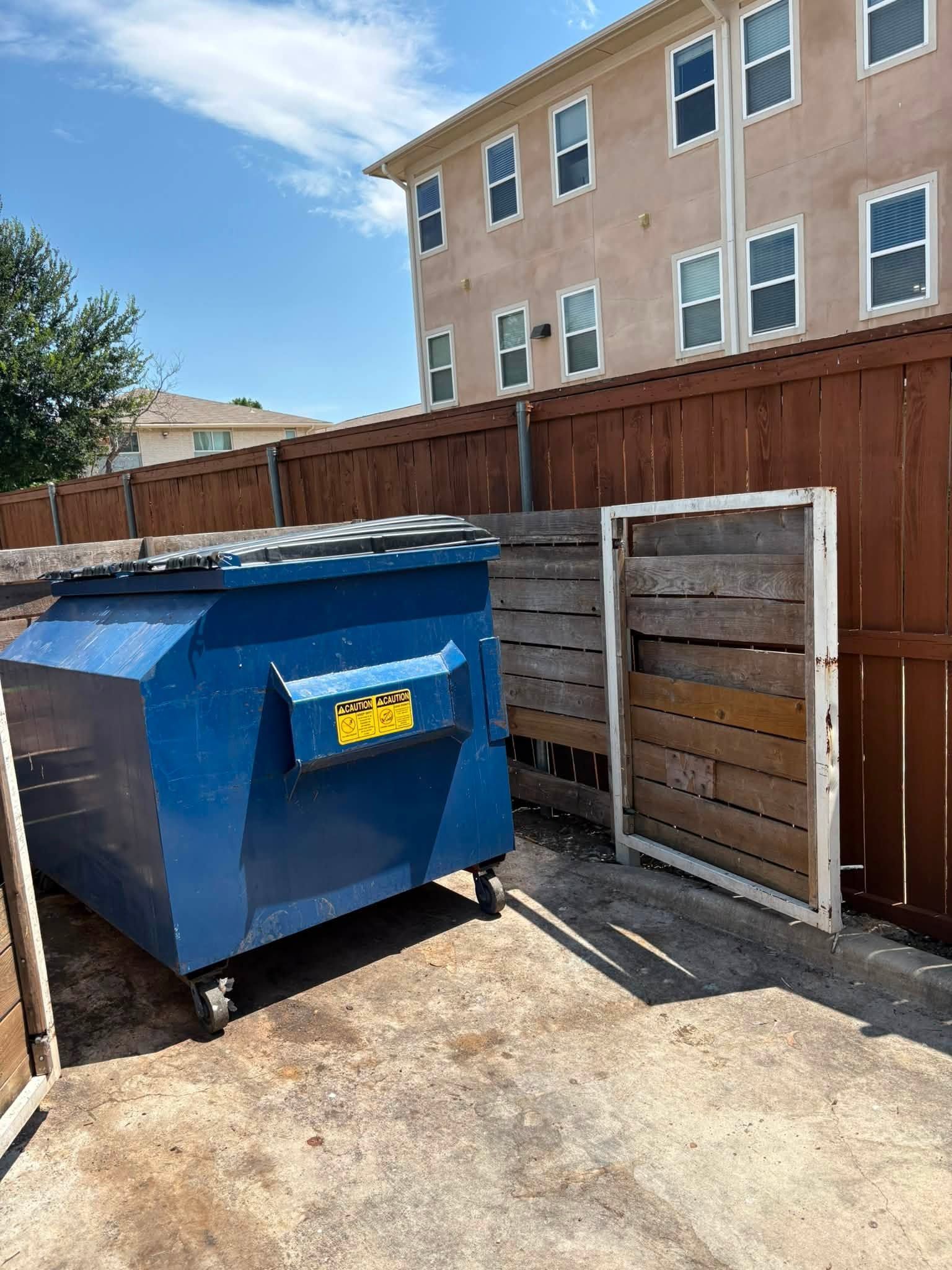 Blue dumpster in a fenced alley beside a beige building under a bright sky.