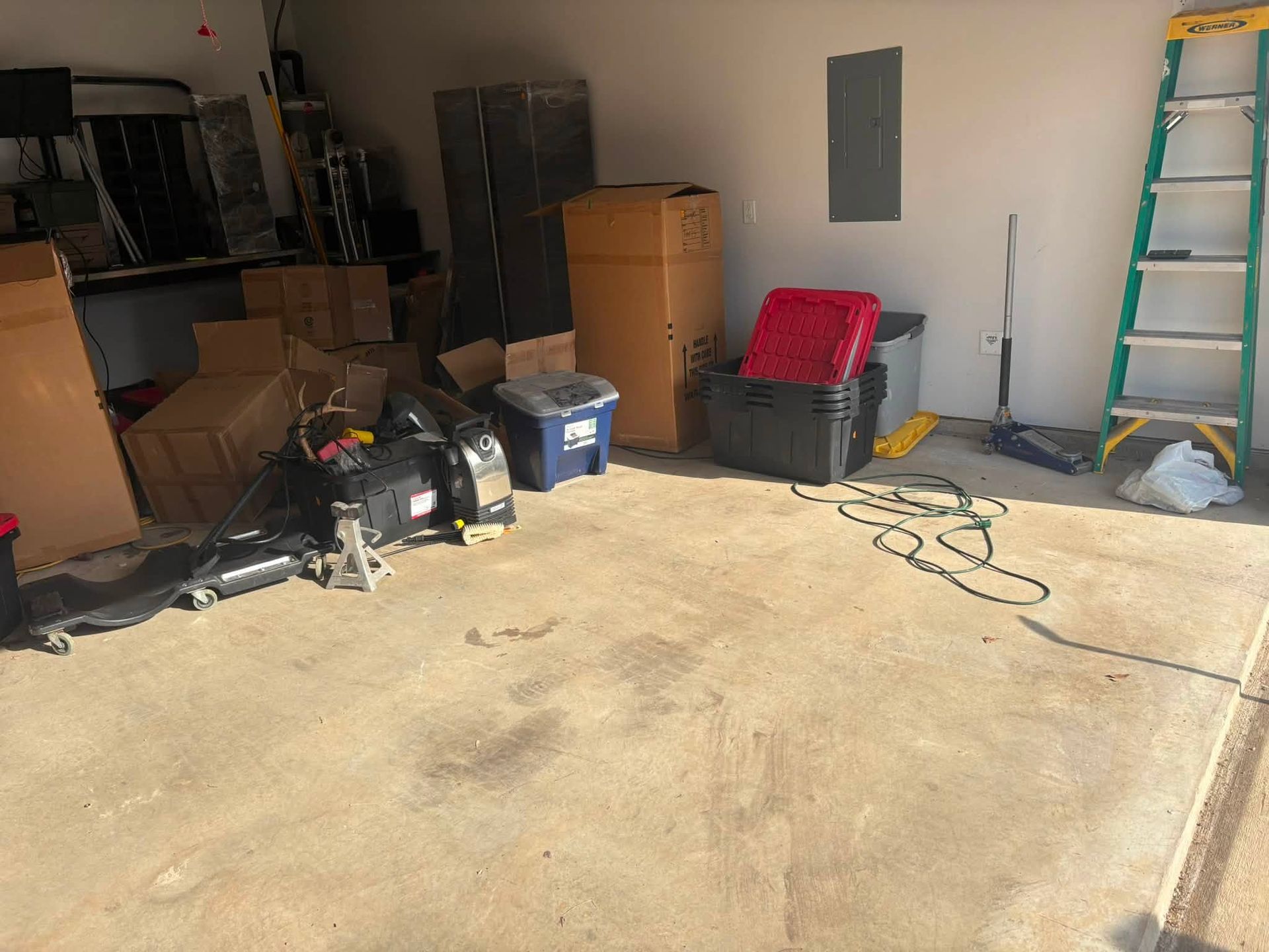 Cluttered garage with boxes, tools, a ladder, and a red chair on a concrete floor.