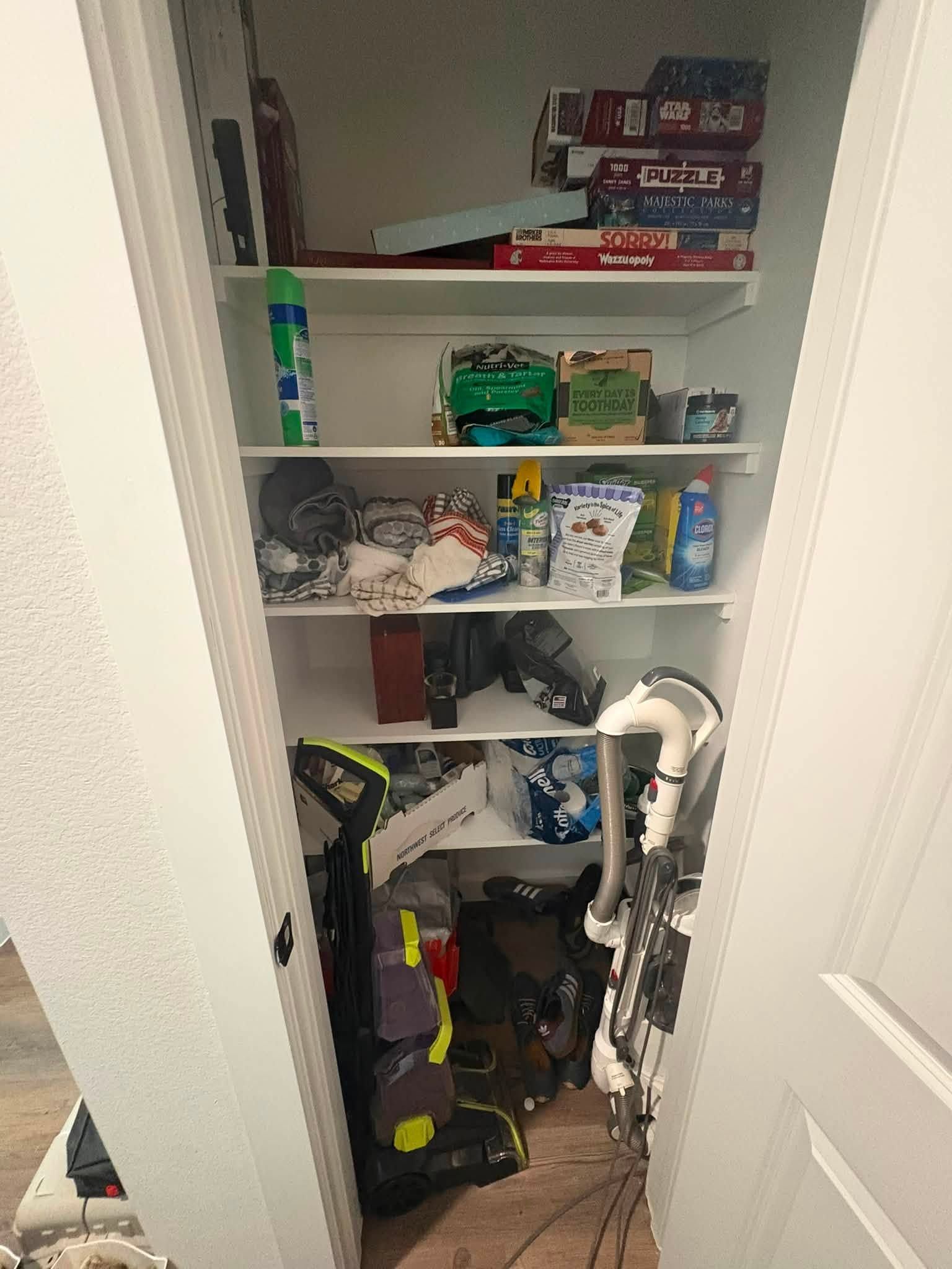 Cluttered pantry with shelves of groceries and household items, plus a vacuum cleaner on the floor.