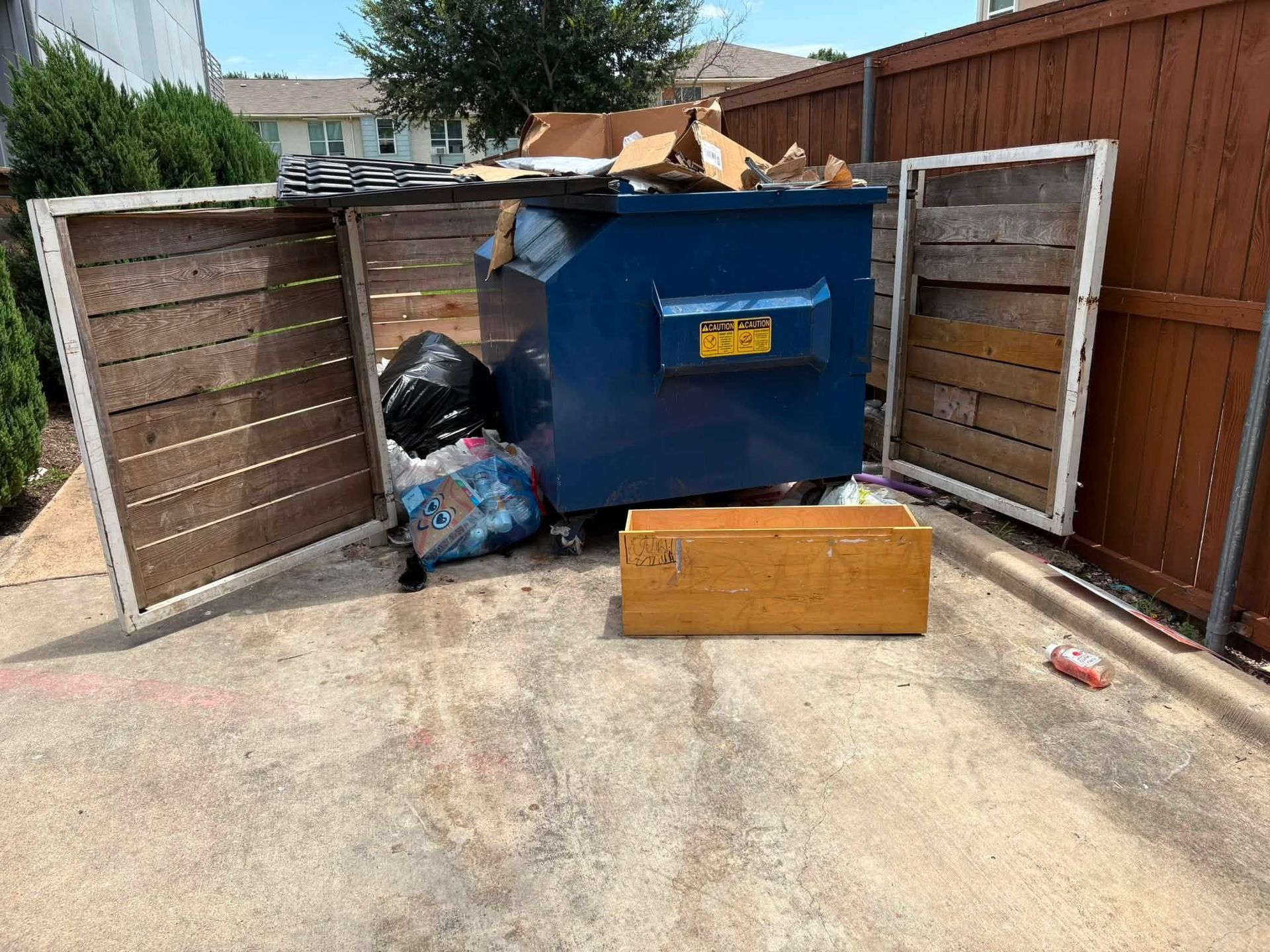 Overflowing blue dumpster with trash beside broken wooden fence in a driveway