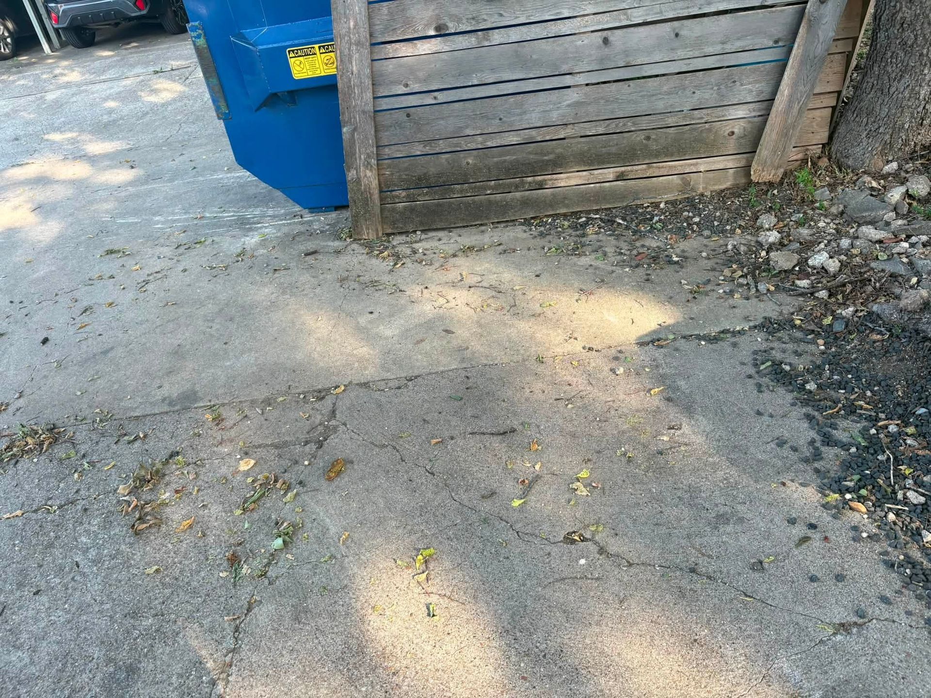 Concrete driveway beside a blue dumpster and wooden fence in bright sunlight.