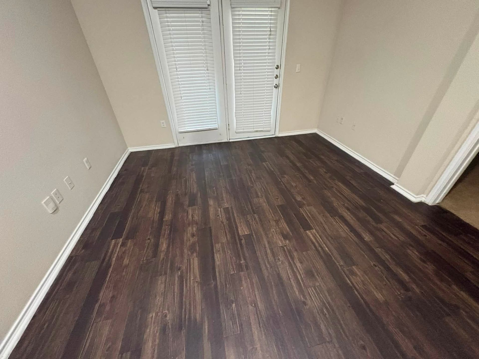 Empty room with dark wood floors and white walls, facing French doors with closed blinds.