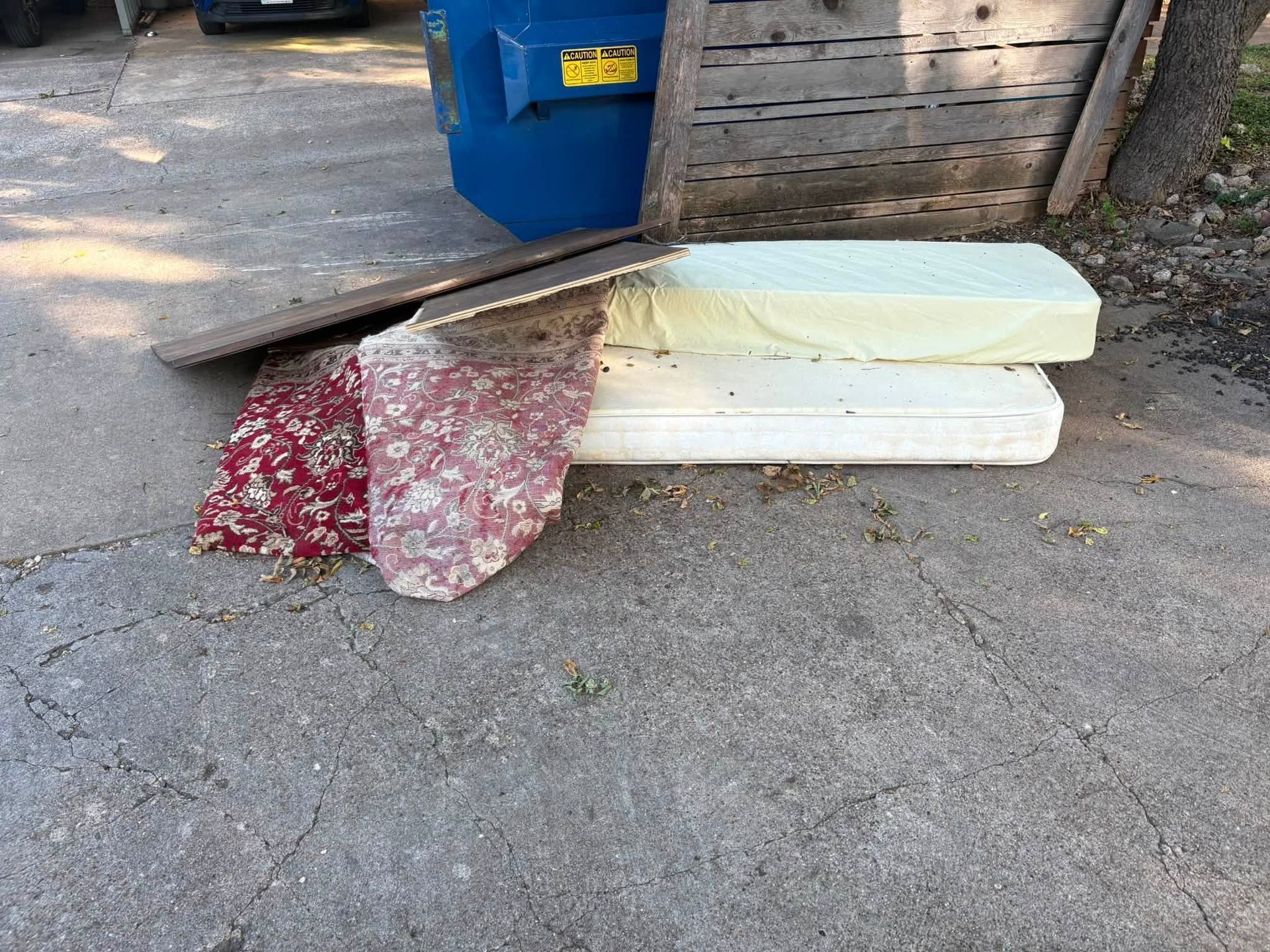 Mattress and plywood piled on a driveway beside a blue recycling bin and wooden shed.