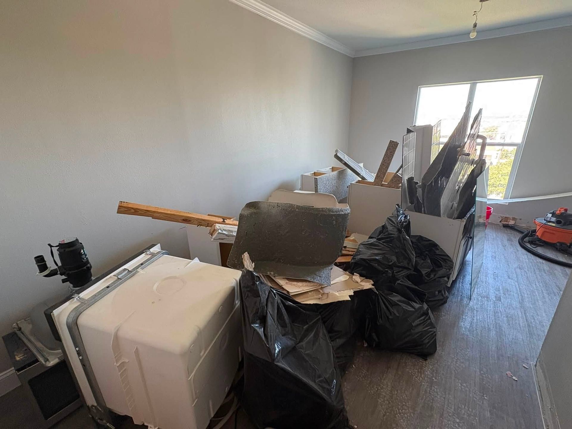 Cluttered room with boxes, bags, and furniture piled near a window on a hardwood floor.