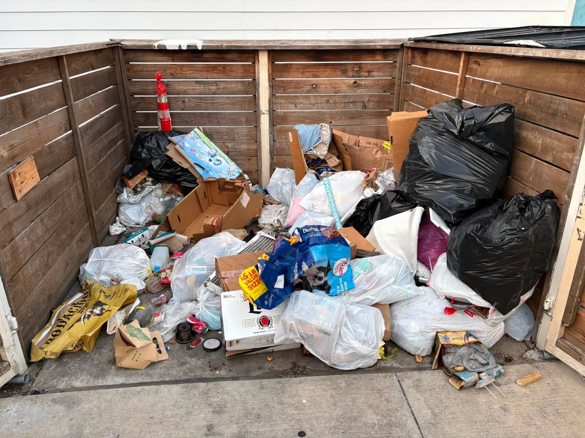 Overflowing trash bags and scattered garbage piled in a fenced outdoor corner