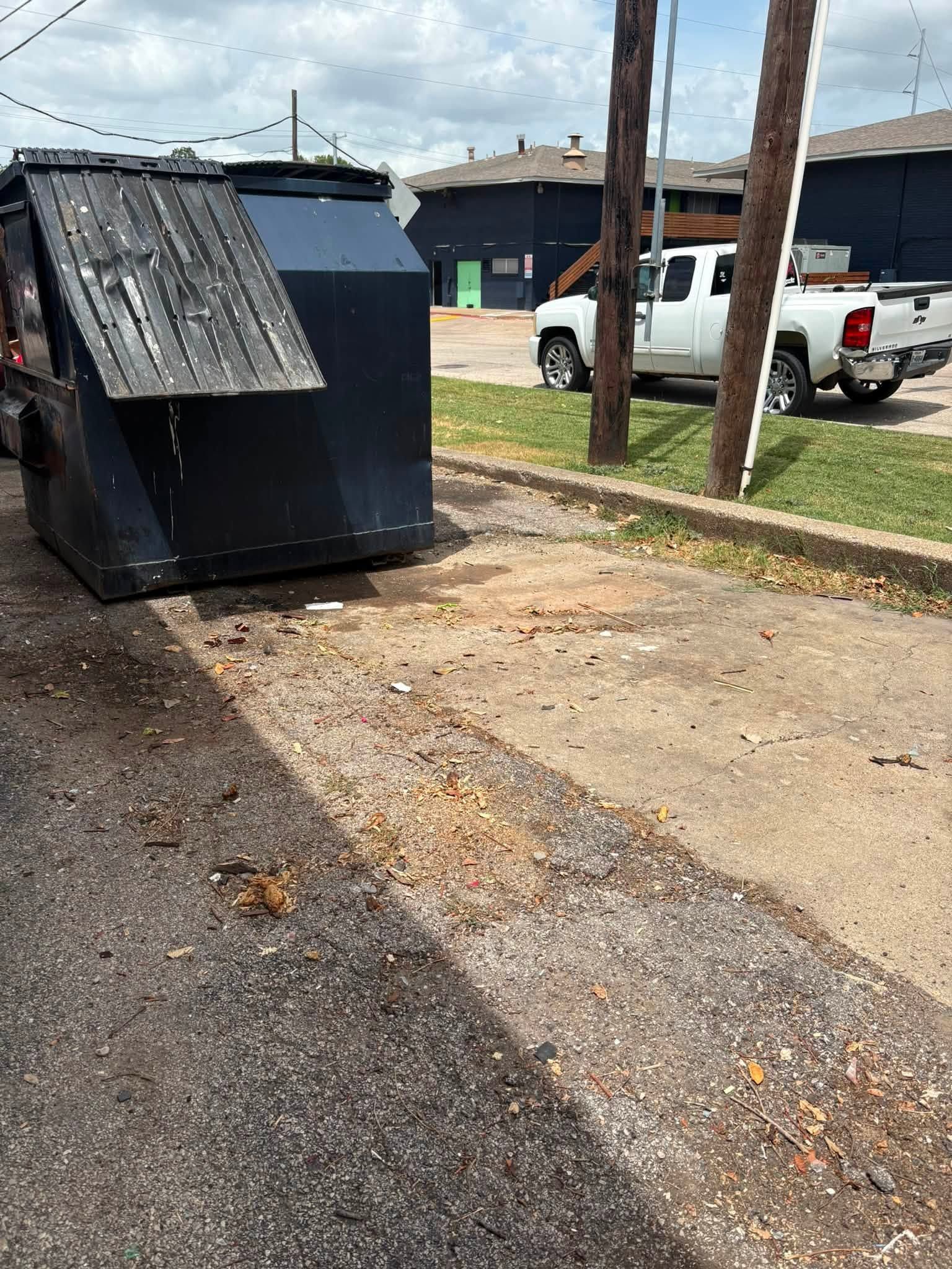 Overturned black dumpster beside a driveway and white pickup truck on a dirt lot.