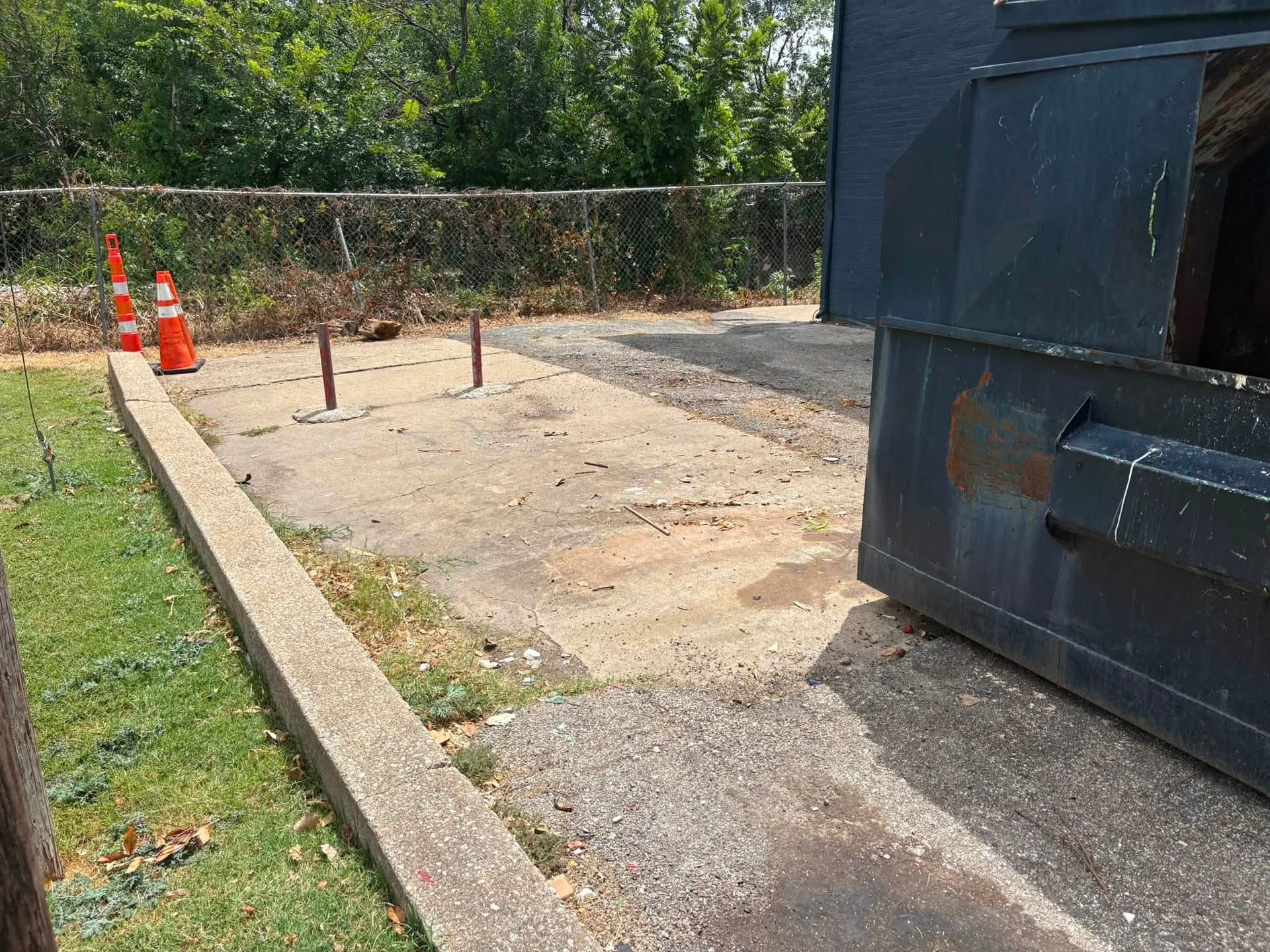 Empty concrete lot beside a large dumpster, with orange cones and a chain-link fence.