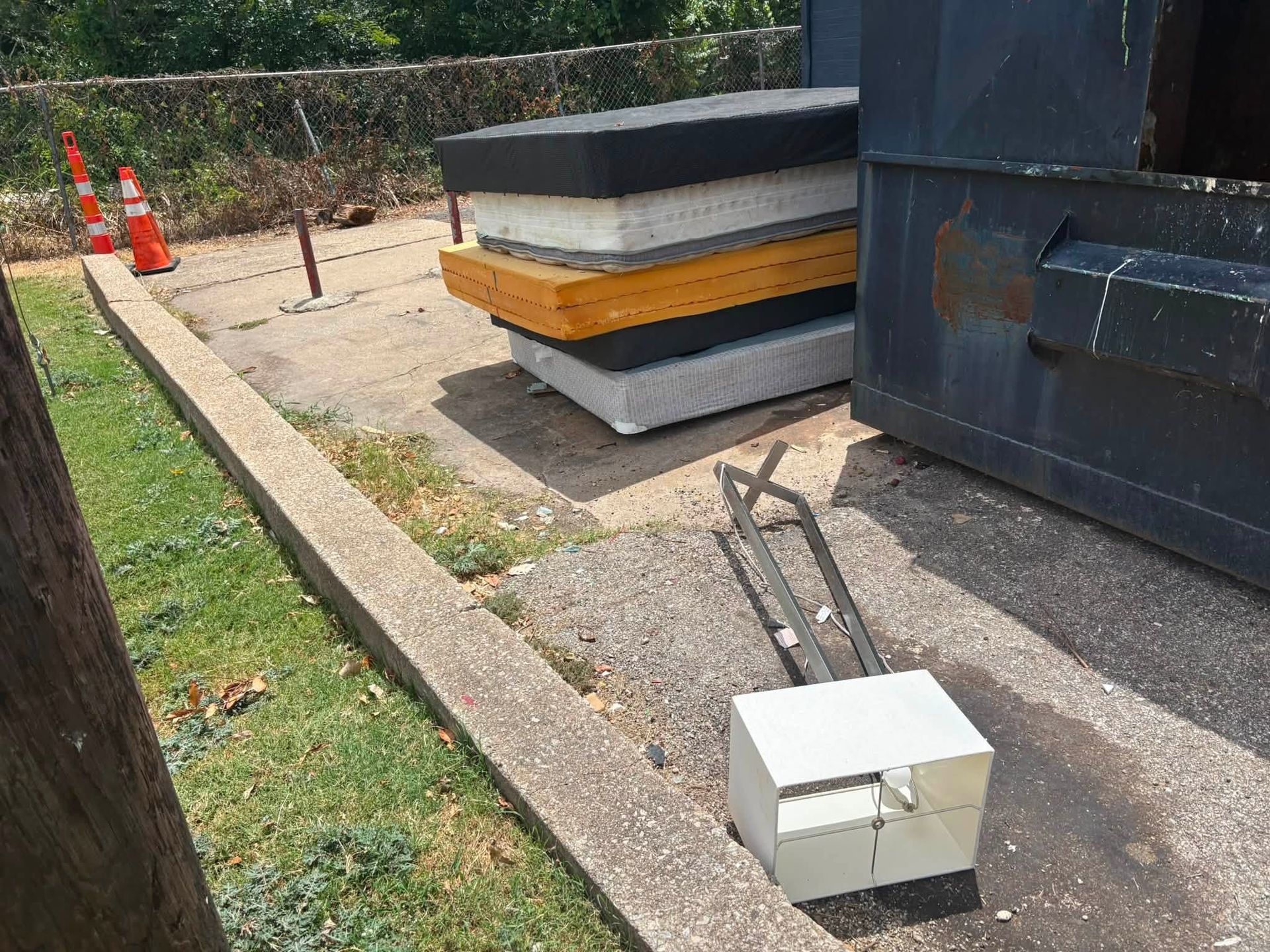 Outdoor construction area with stacked concrete barriers, cones, a metal cart, and a white box on pavement.