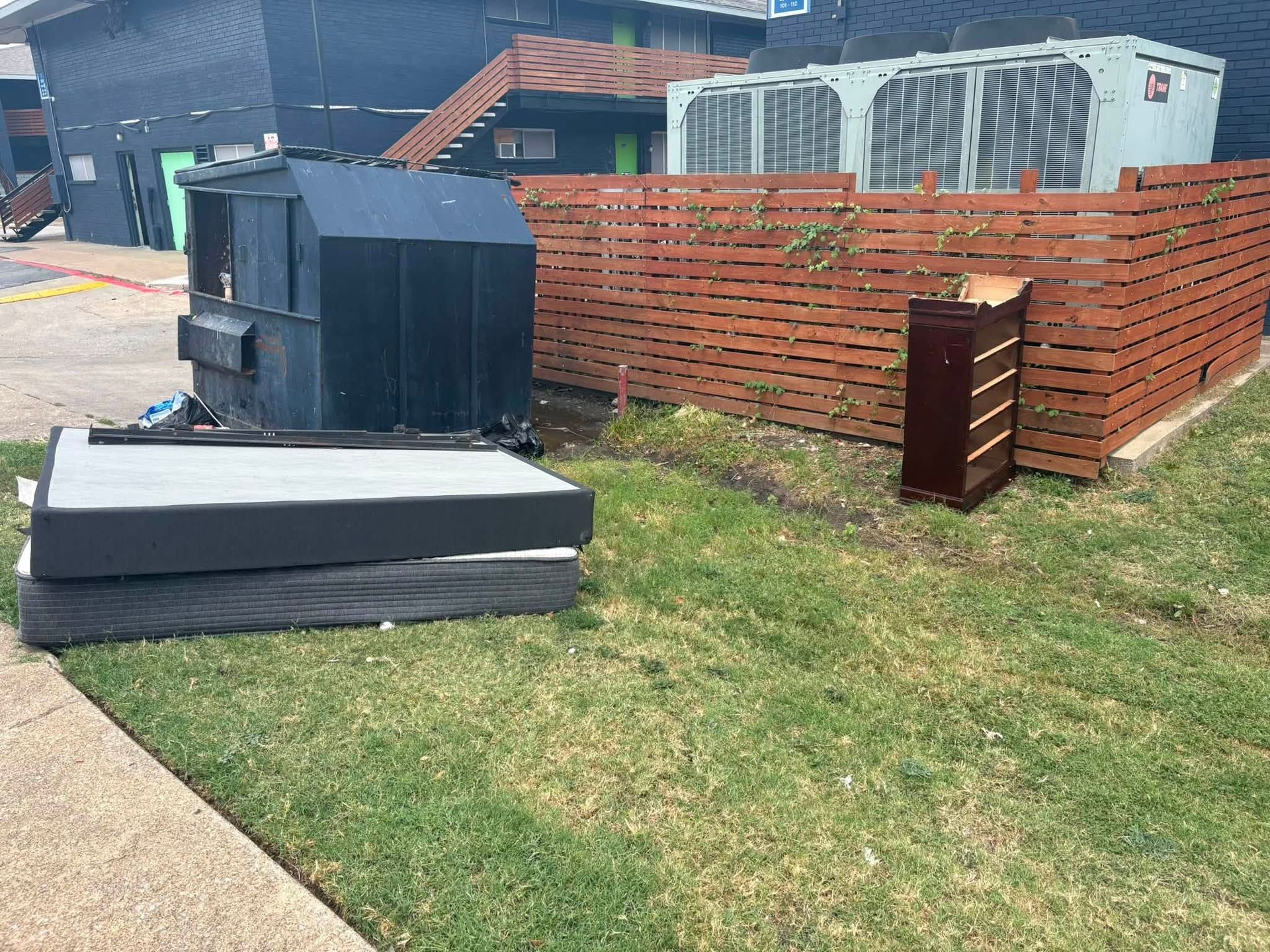 Outdoors near a parking lot, stacked mattresses and a black appliance sit by a brick wall and grassy verge.