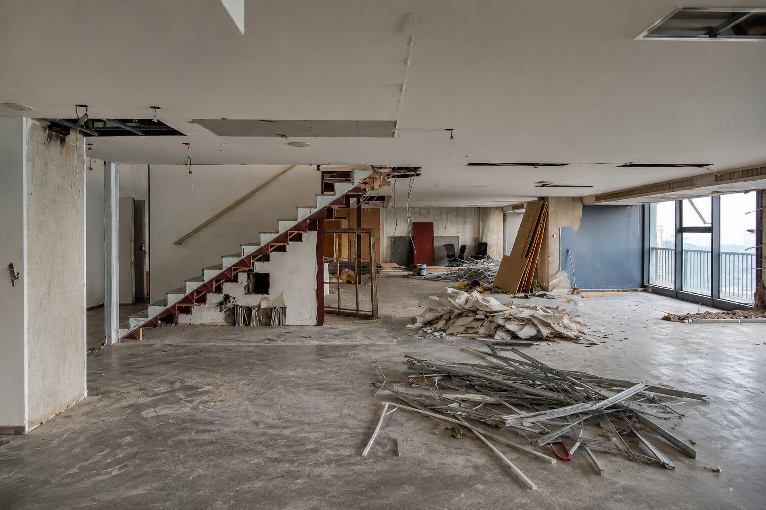 Abandoned interior under renovation with debris on the floor, exposed staircase, and large windows.