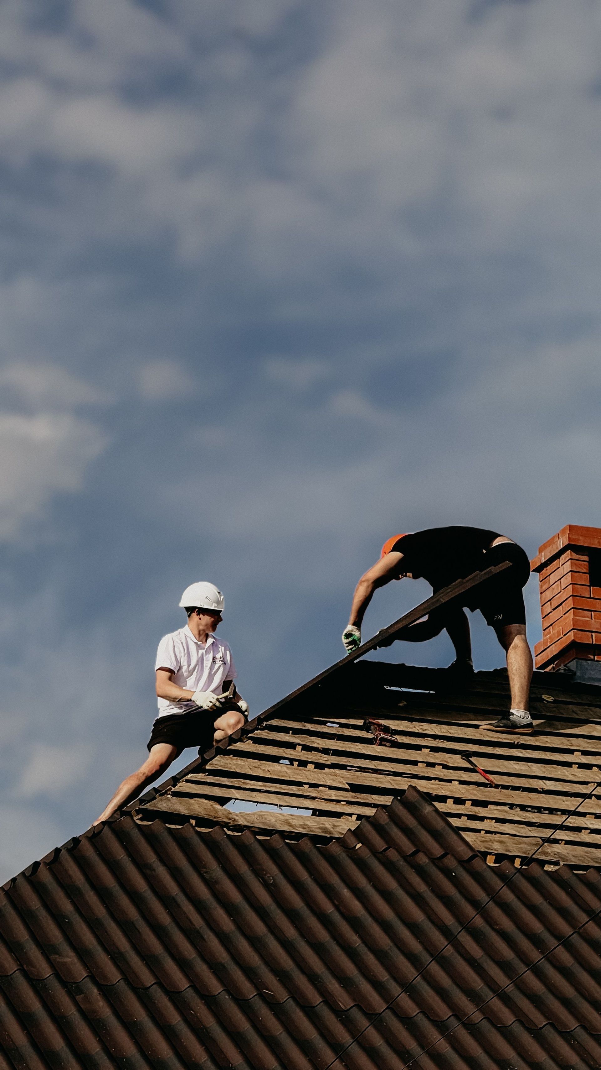 Two roofers on a shingled roof, one bending over near a brick chimney under a cloudy sky