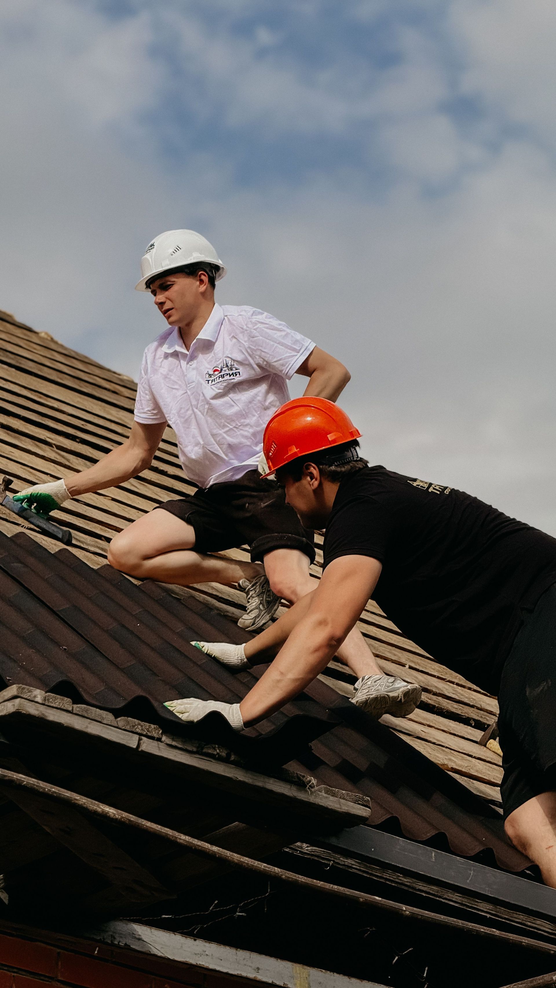 Two roofers wearing helmets work on a sloped shingle roof under a cloudy sky.