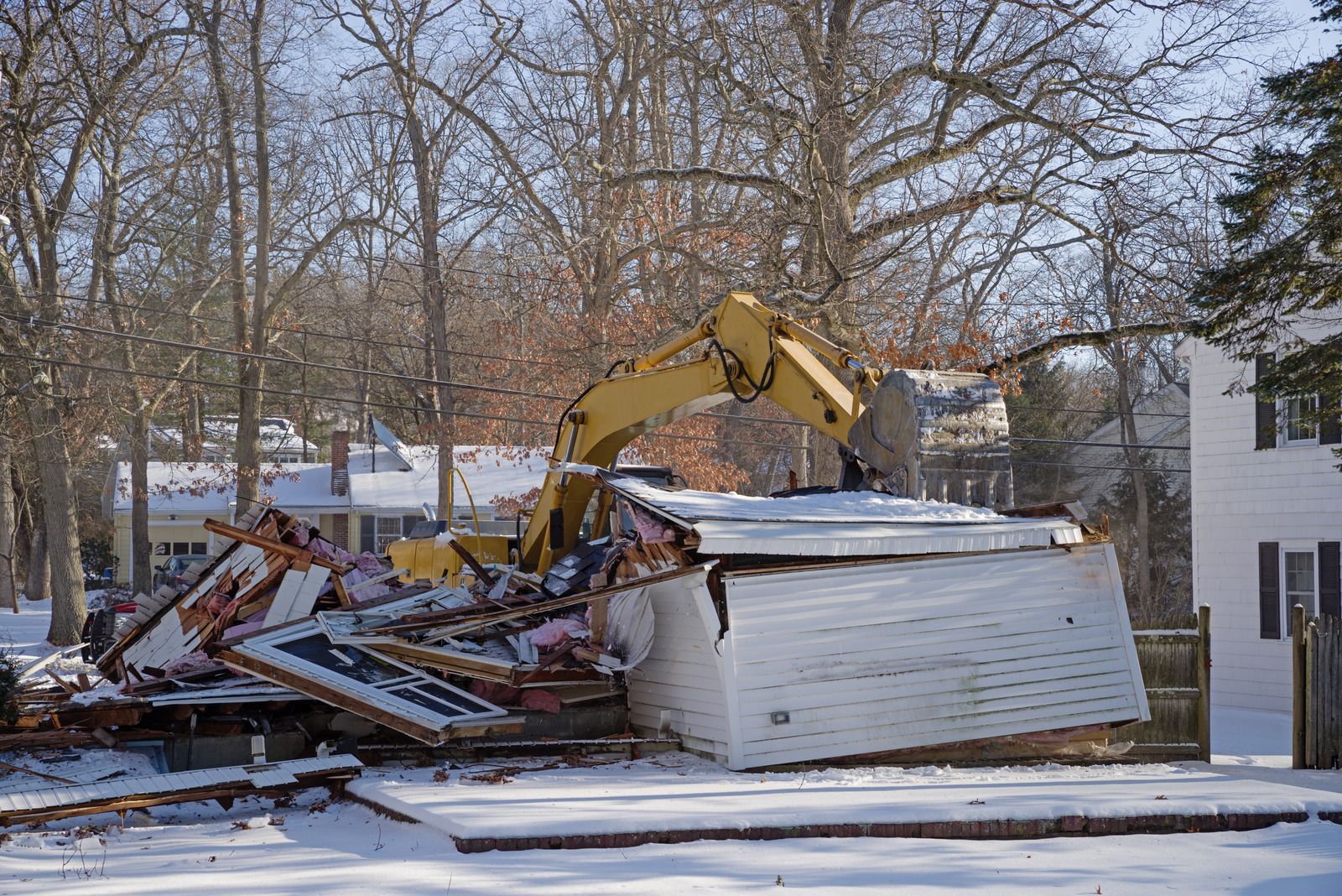 Excavator demolishing a collapsed white house in a snowy residential yard with bare trees nearby