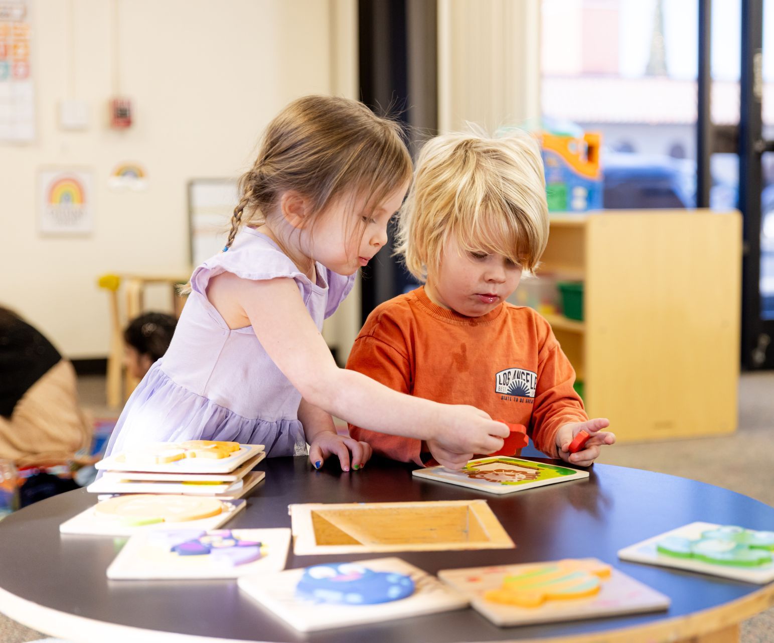 Two children playing with puzzles at a table in a classroom.