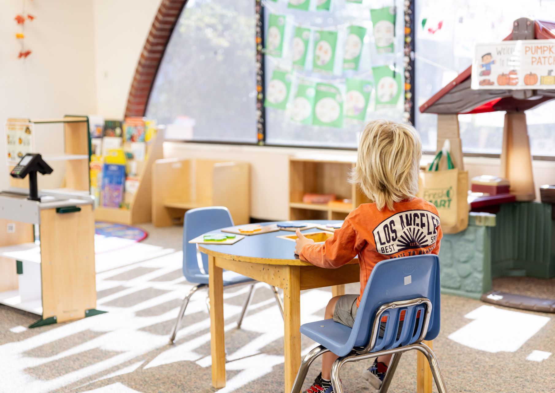 A child with blonde hair wearing an orange long-sleeve shirt sits at a small wooden table in a brightly lit classroom.
