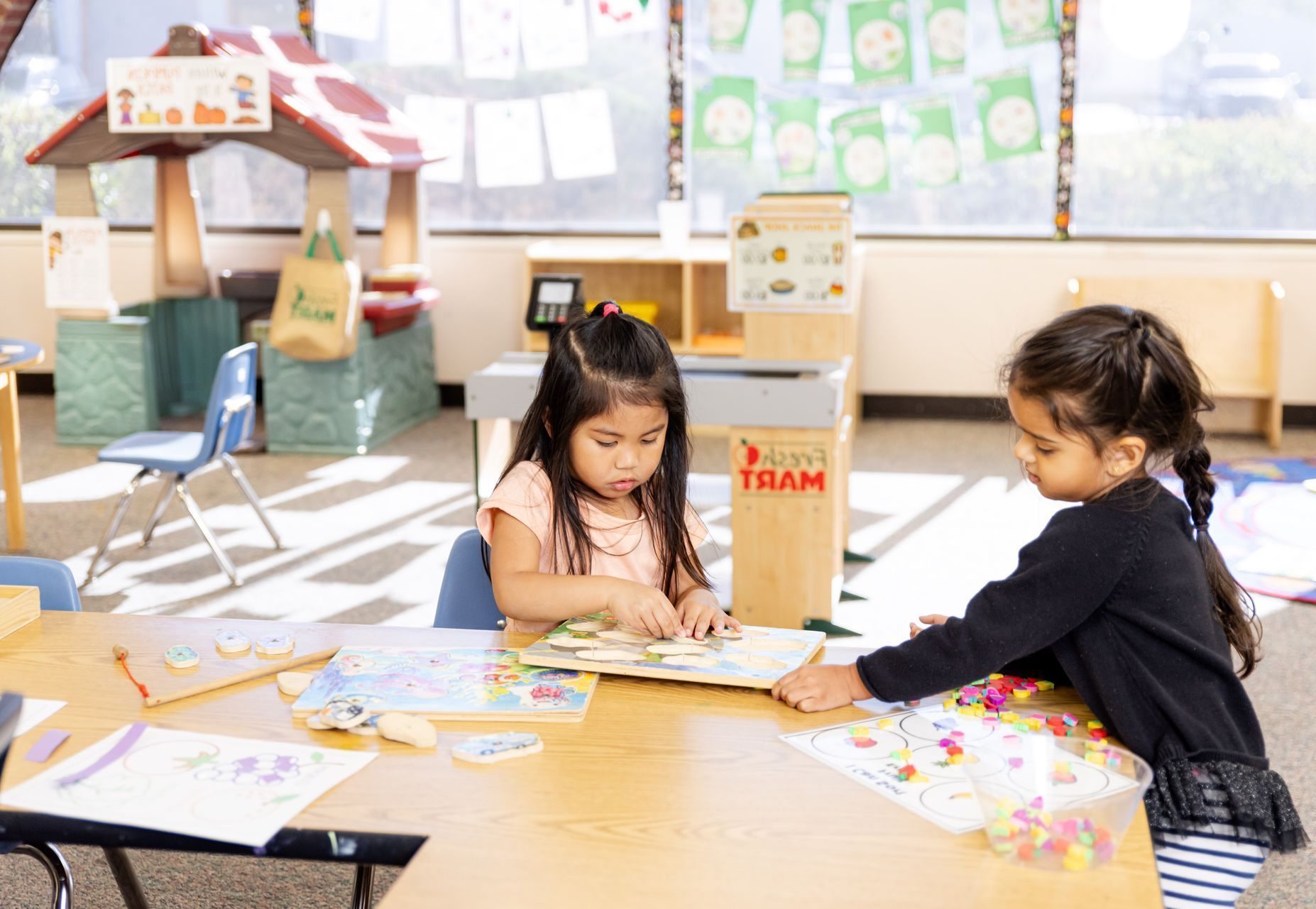 Children sitting at a classroom table, drawing and working on papers in a bright classroom
