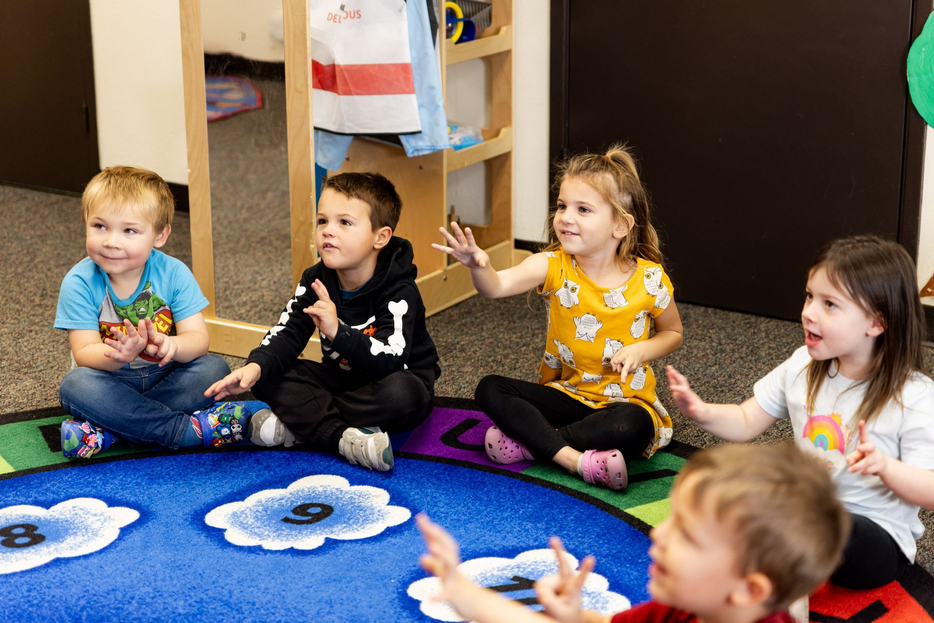 Children sitting on a colorful rug in a classroom, clapping and playing together