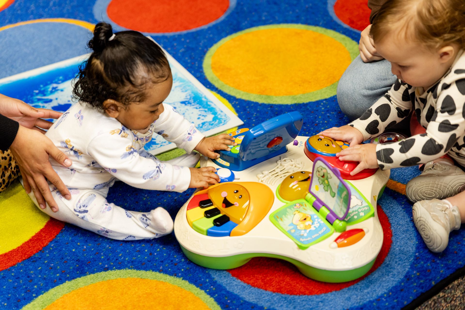 Two toddlers play with a musical toy on a colorful rug. One has a caregiver's hand on them.