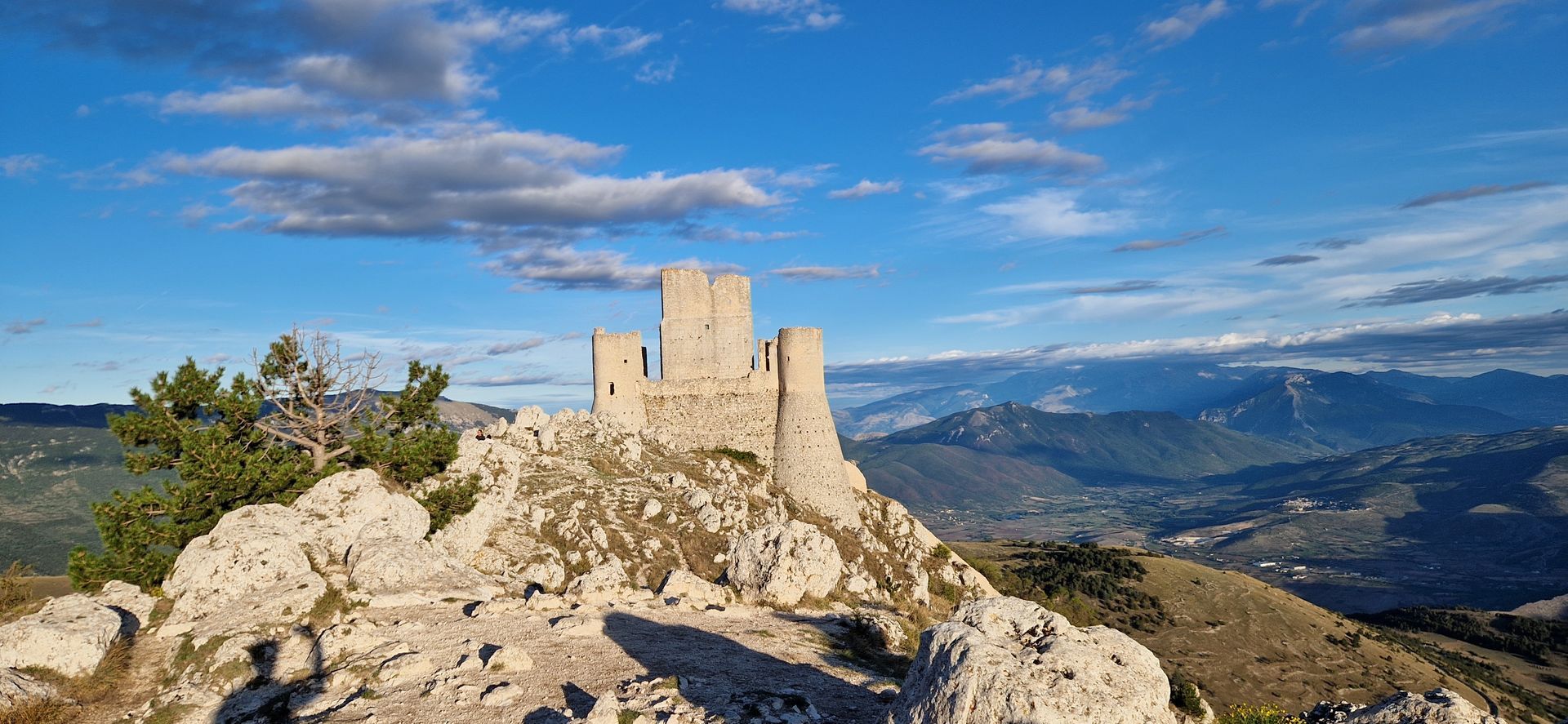 le torri di castello si ergono sulla cima di una montagna scoscesa e rocciosa, sotto un cielo azzurro con nuvole sparse.