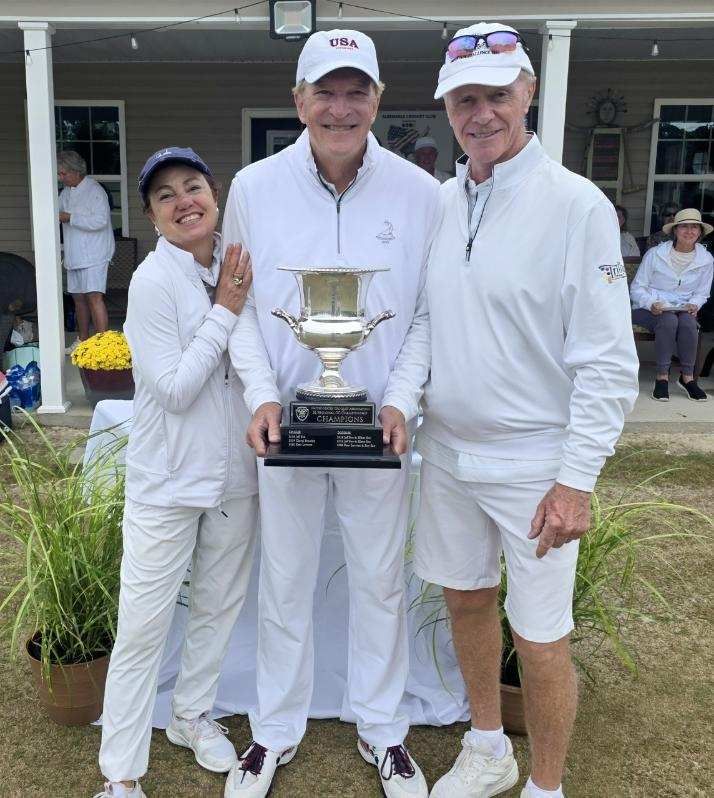 Three people in white outfits pose with a trophy outdoors. Woman on left smiles. Man in center holds trophy. Man on right smiles.