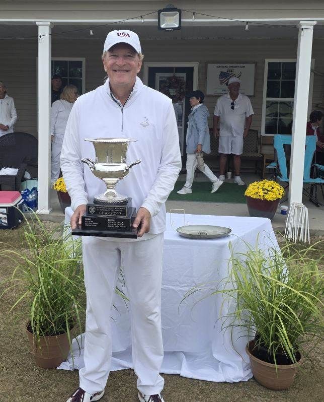 Man in white holding a trophy, standing near a table with flowers. Outdoors in front of a building.