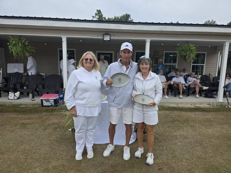 Three people holding trophies outside a building; all wearing white. Man in the center is smiling.