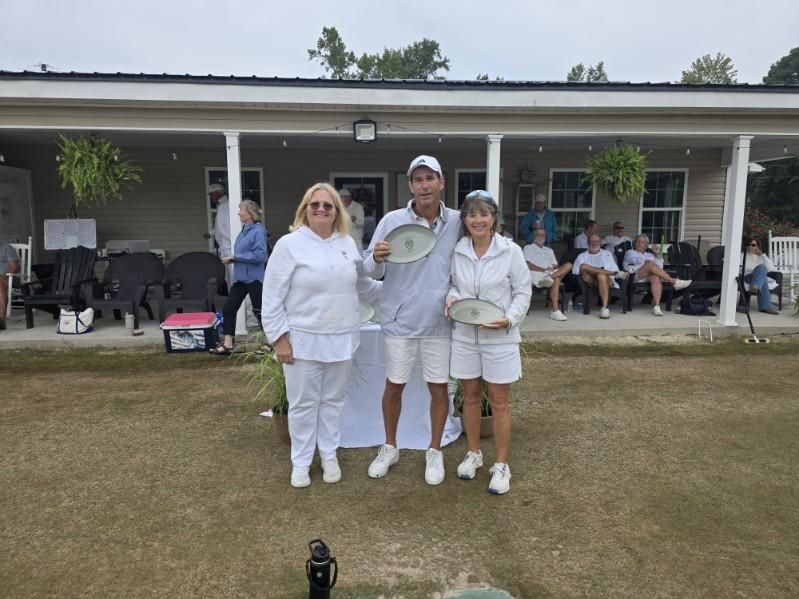 Three people in white holding trophies stand on grass; people watch in the background near a building.
