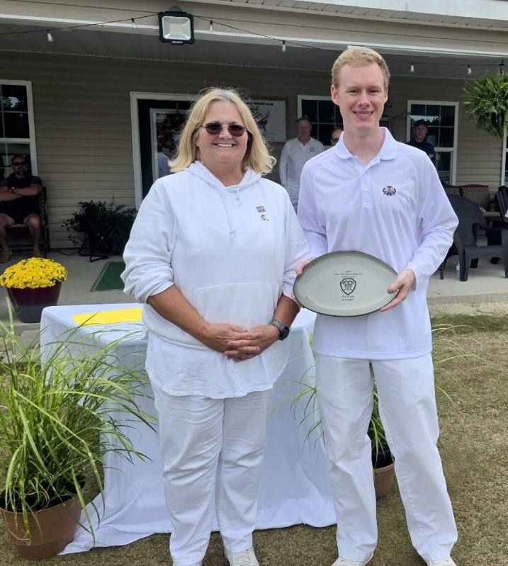 Woman and man in white clothing holding a dish, smiling, outdoors near a building.