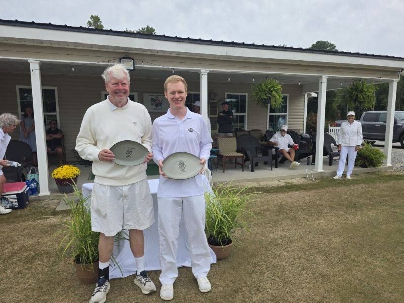 Two people holding trophies stand outside a building with others nearby; the setting is outdoors.