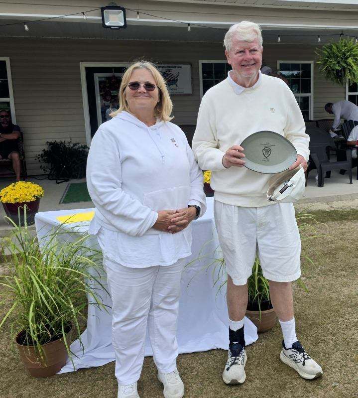 Woman and man in white clothing stand outdoors. Man holds a trophy.