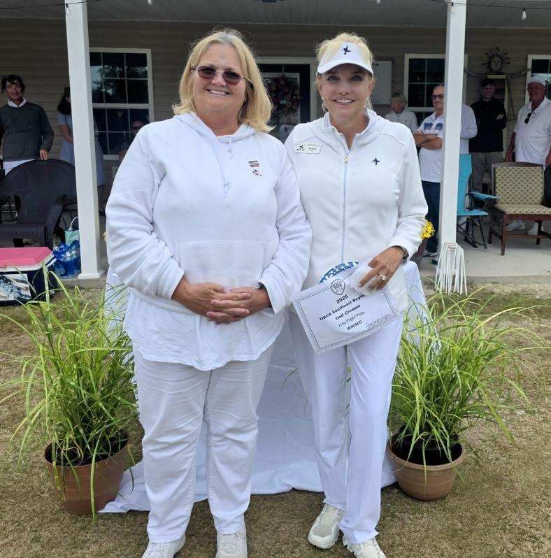 Two women pose outdoors, one holding a certificate. They are in white clothing. Plants in pots flank them.