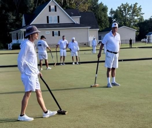 People playing croquet on a green lawn. Players wear white and use mallets near a large house.