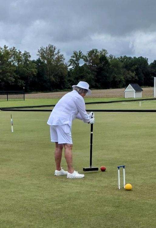 Person in white playing croquet on a green lawn, holding mallet, with white hat, and a cloudy sky.