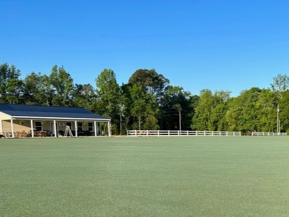 A green lawn with a white fence, building, and trees against a bright blue sky.