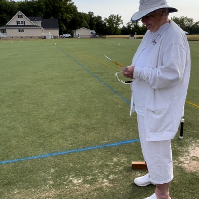 Person in white outfit and hat on a croquet court, looking at papers.