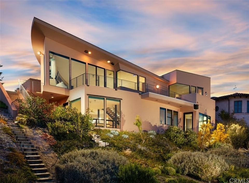 Modern two-story house with a sloped roof and large windows, situated on a grassy hillside at sunset.