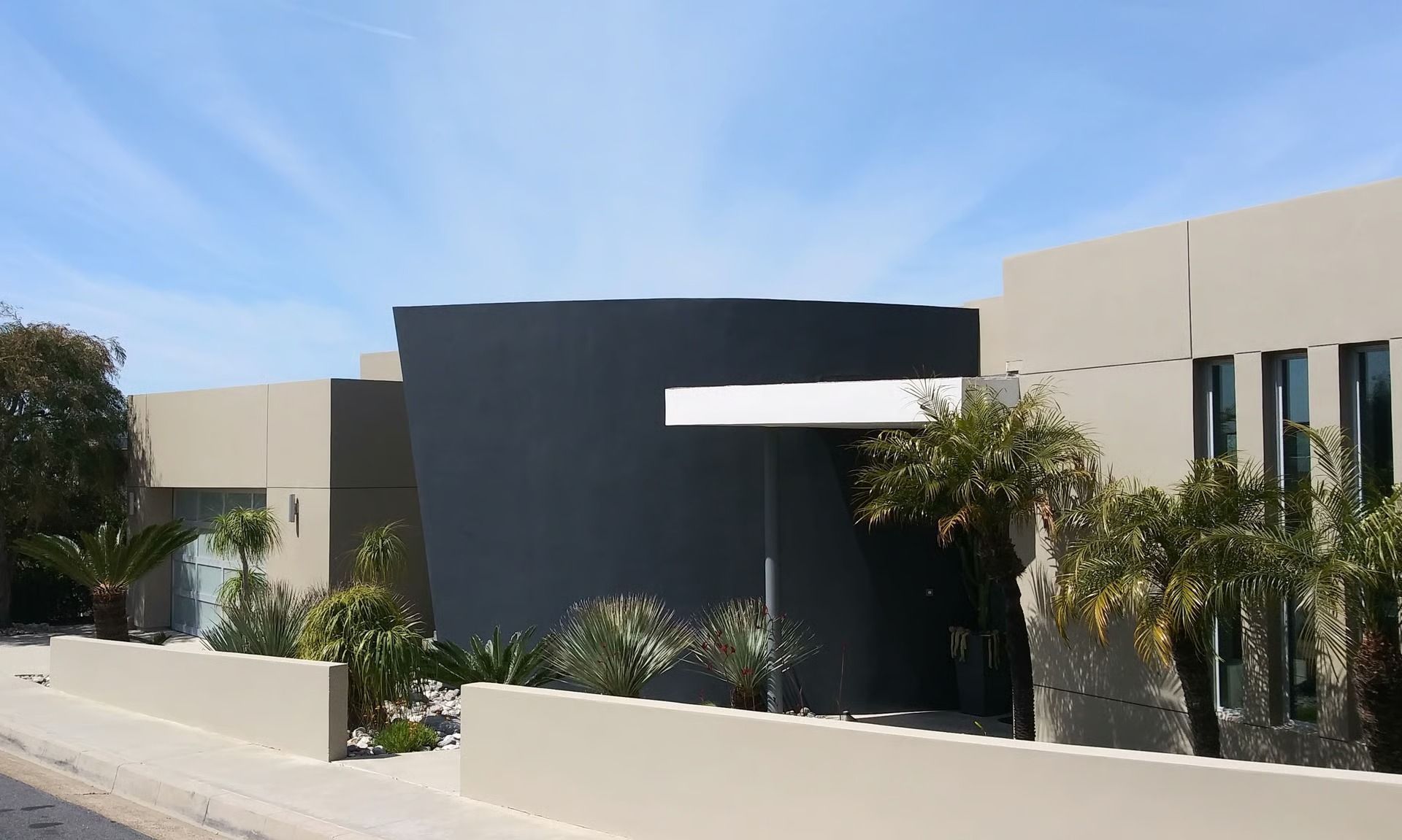 Modern house with beige walls, a dark grey focal section, a flat roofline, and landscaped shrubs behind a low wall.
