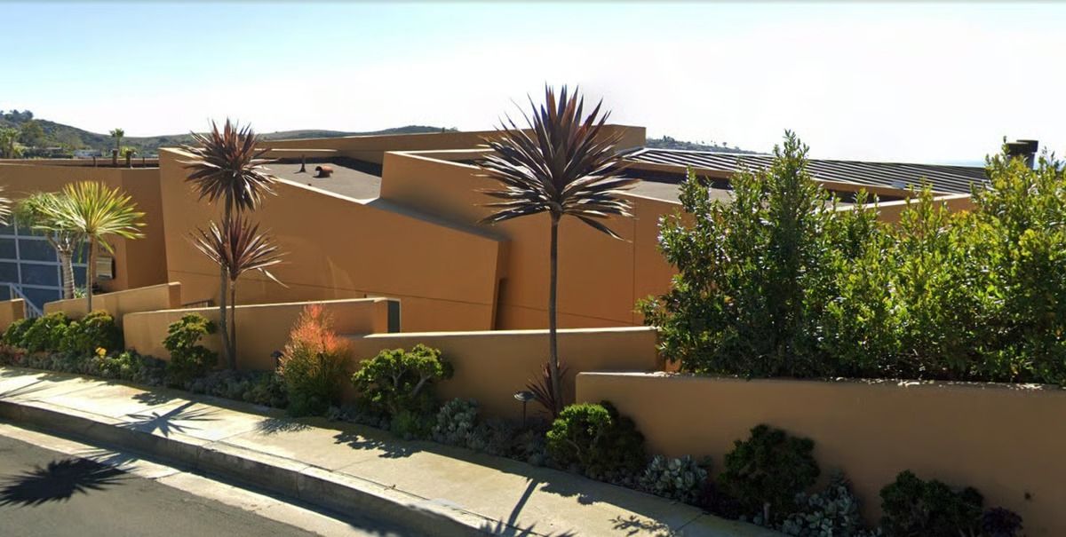 A modern home with an angular, tan exterior, a low wall, and landscaping along a sunny street.