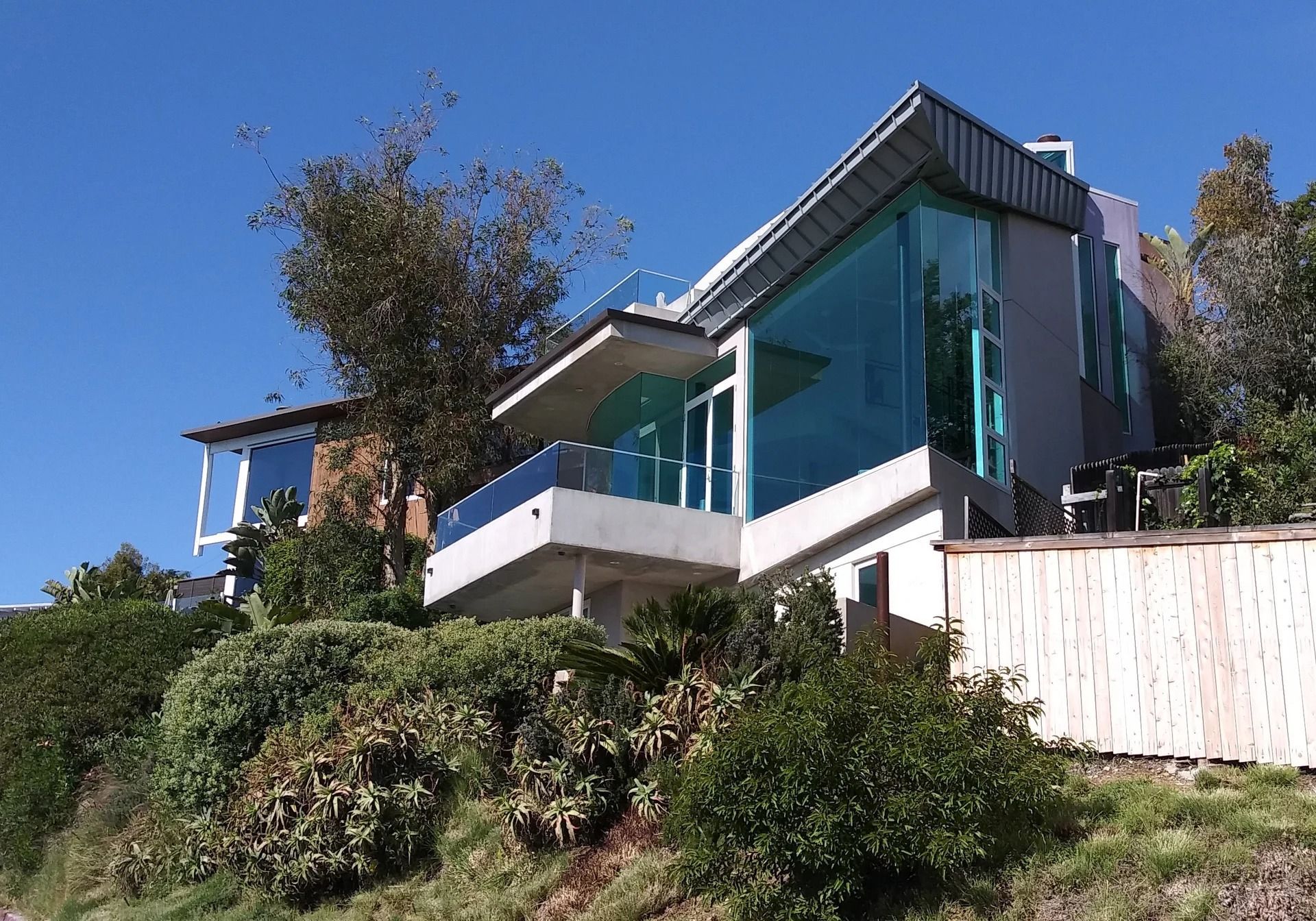 A modern, multi-story house with large glass windows and balconies, perched on a grassy hillside under a clear blue sky.
