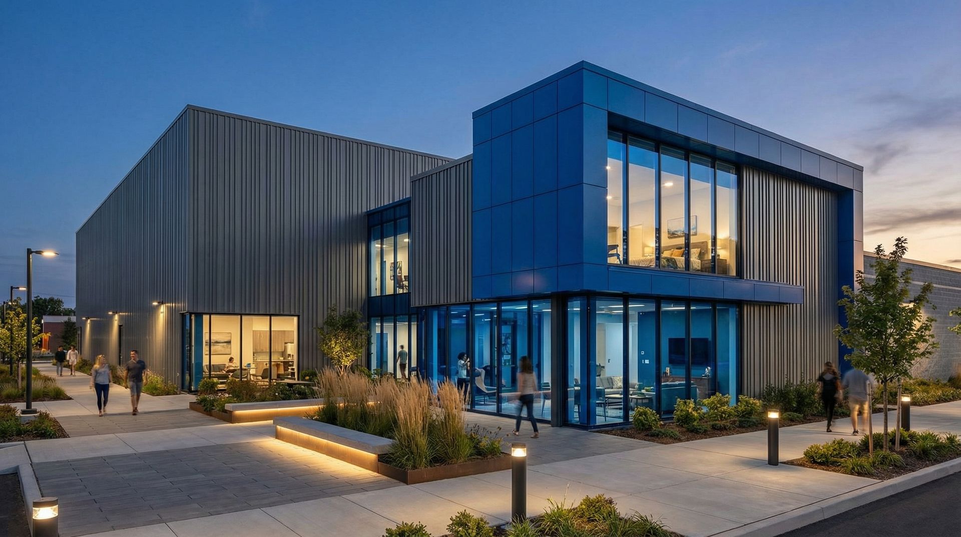 Modern building exterior at twilight with blue metal panels, glass windows, illuminated landscaping, and pedestrians.