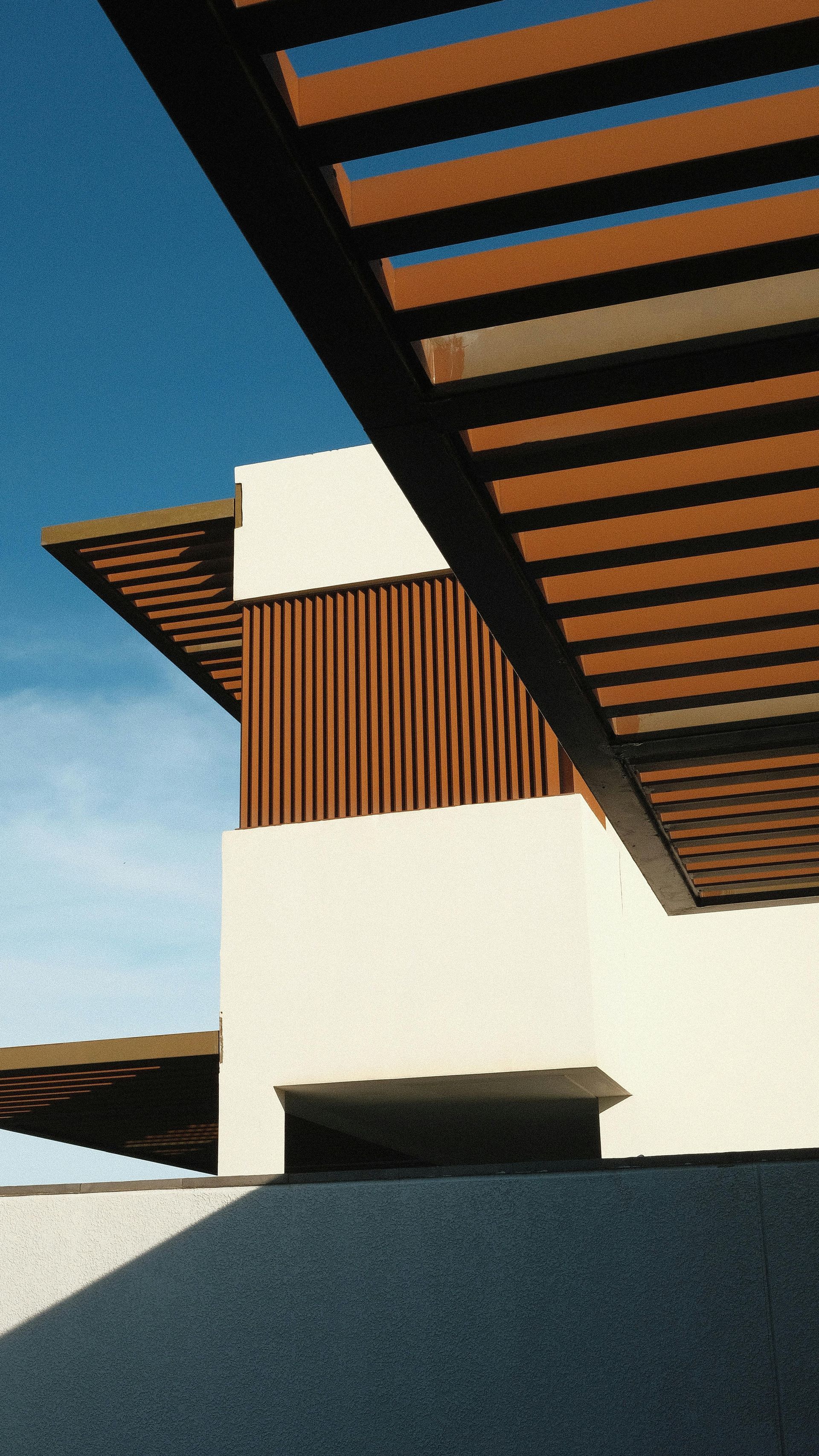 Low-angle shot of a modern building with white walls, wood-slat siding, and a wooden pergola against a bright blue sky.