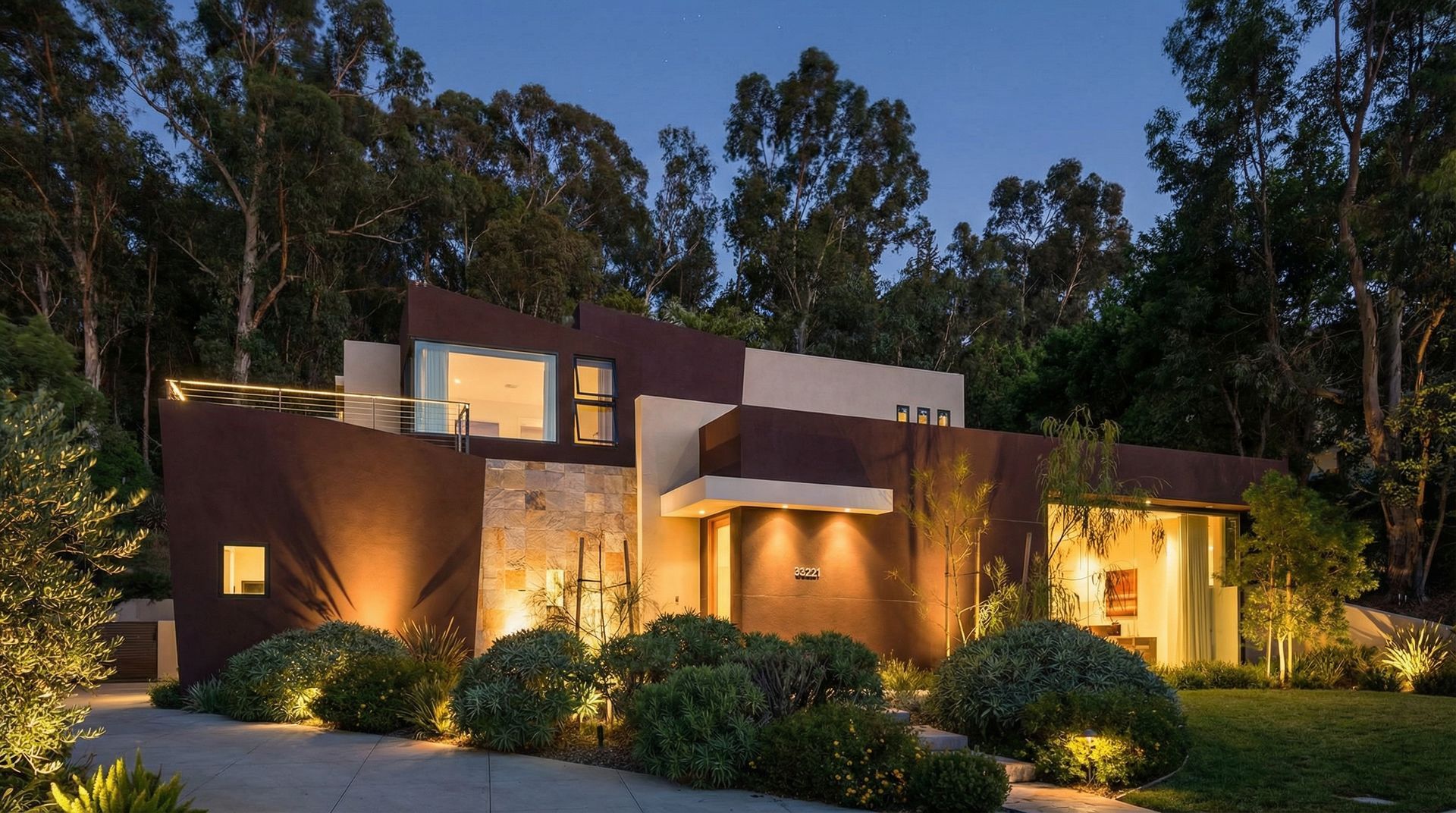 A modern, multi-level home with dark brown and beige accents is illuminated by warm evening lights amidst lush trees.