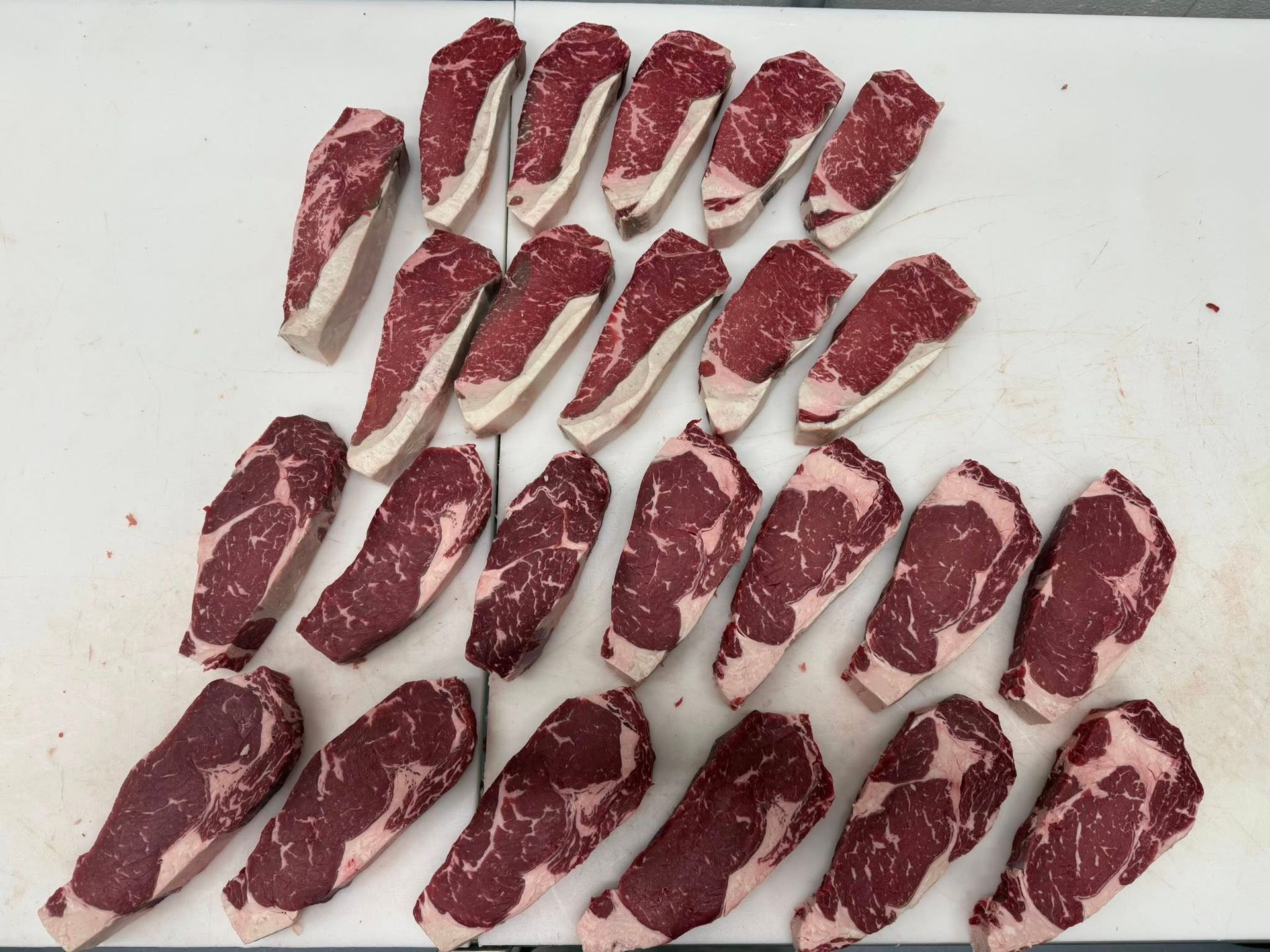 Raw steaks arranged on a white surface, varying in size, red meat with white fat around the edges.